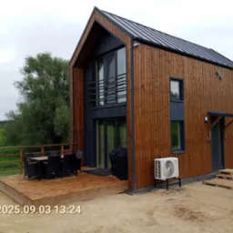 Nowoczesny dom modułowy z drewnianą elewacją, tarasem i klimatyzacją. Widok z zewnątrz w pochmurny dzień. Na pierwszym planie piasek i data na zdjęciu.