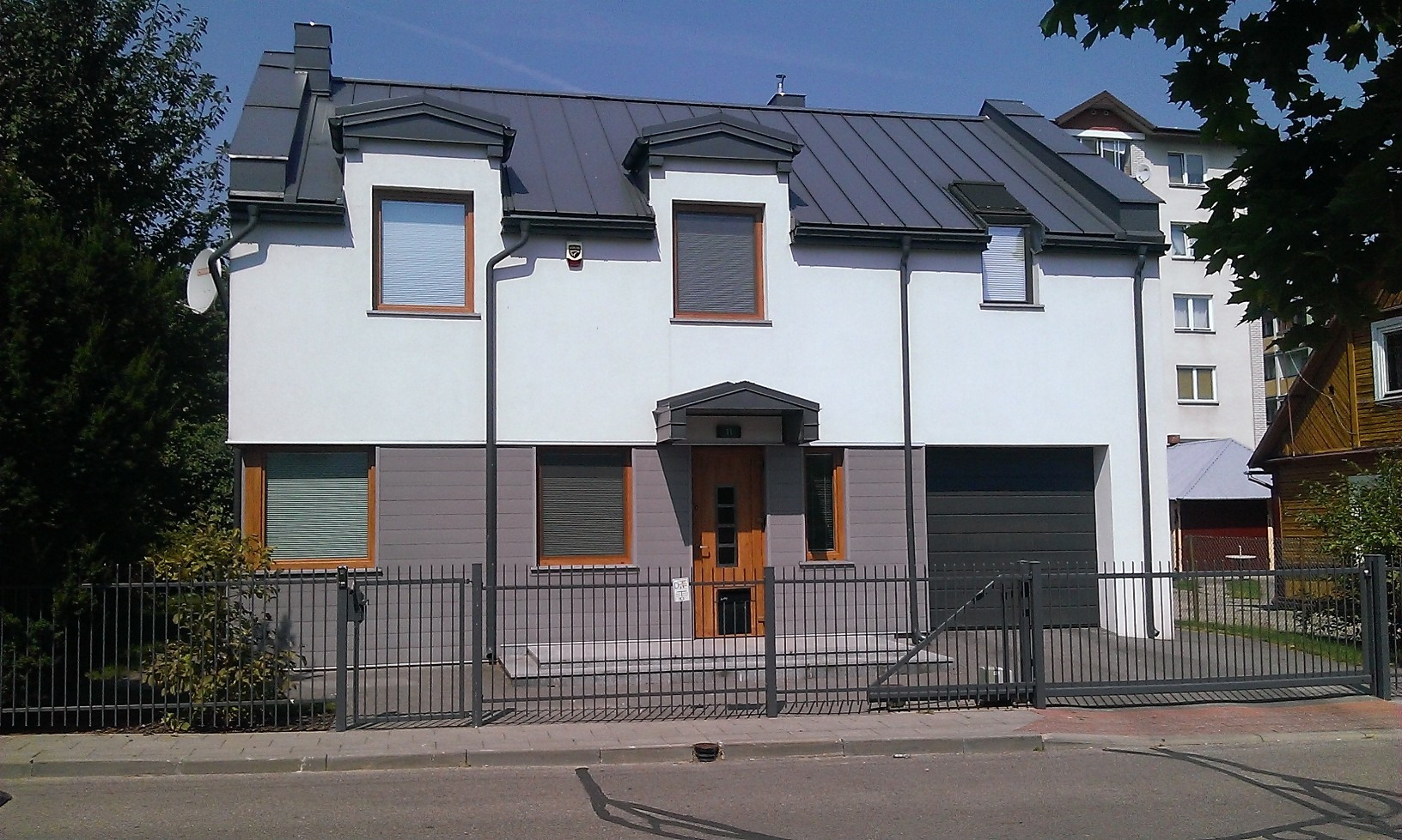 Dwukondygnacyjny dom z szarą, metalową bramą wjazdową i dachem z lukarnami, widziany z poziomu ulicy w słoneczny dzień. Widoczne elementy wykończenia i detale architektoniczne.