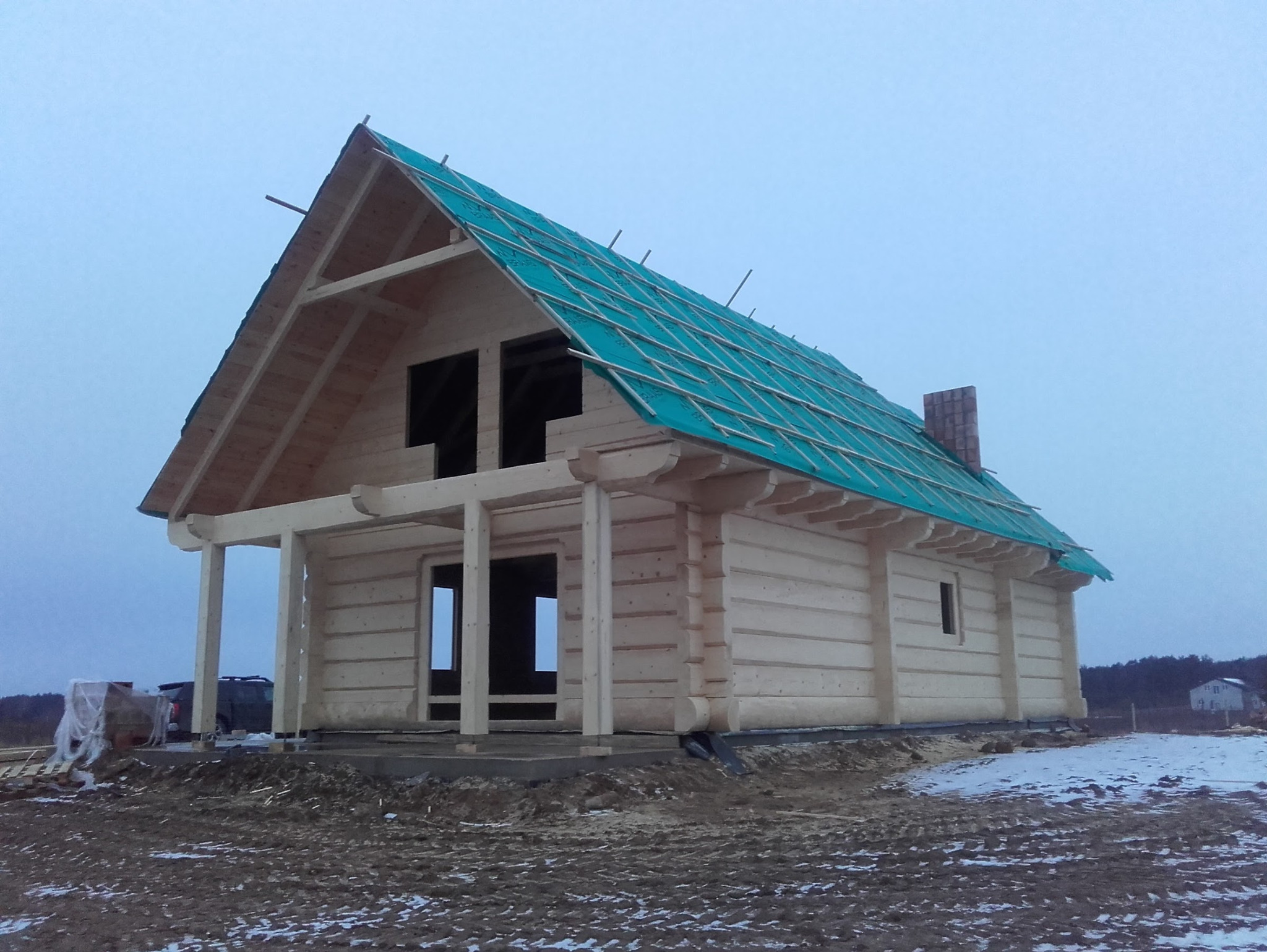 Surowy stan domu z bali, z zieloną membraną na dachu i kominem, na tle zimowego krajobrazu i pochmurnego nieba. Widoczne elementy konstrukcyjne.