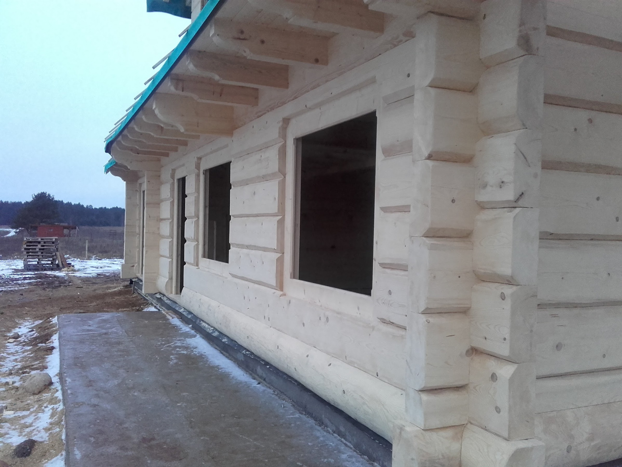 Surowy stan domu z bali: widok z boku na ściany z otworami okiennymi, zadaszenie z zieloną folią i resztki śniegu na ziemi. Widoczne detale łączeń bali.