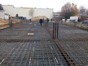 Zbrojenie fundamentu domu jednorodzinnego z widocznymi prętami pionowymi, przygotowane do zalania betonem. W tle budynki i drzewa.