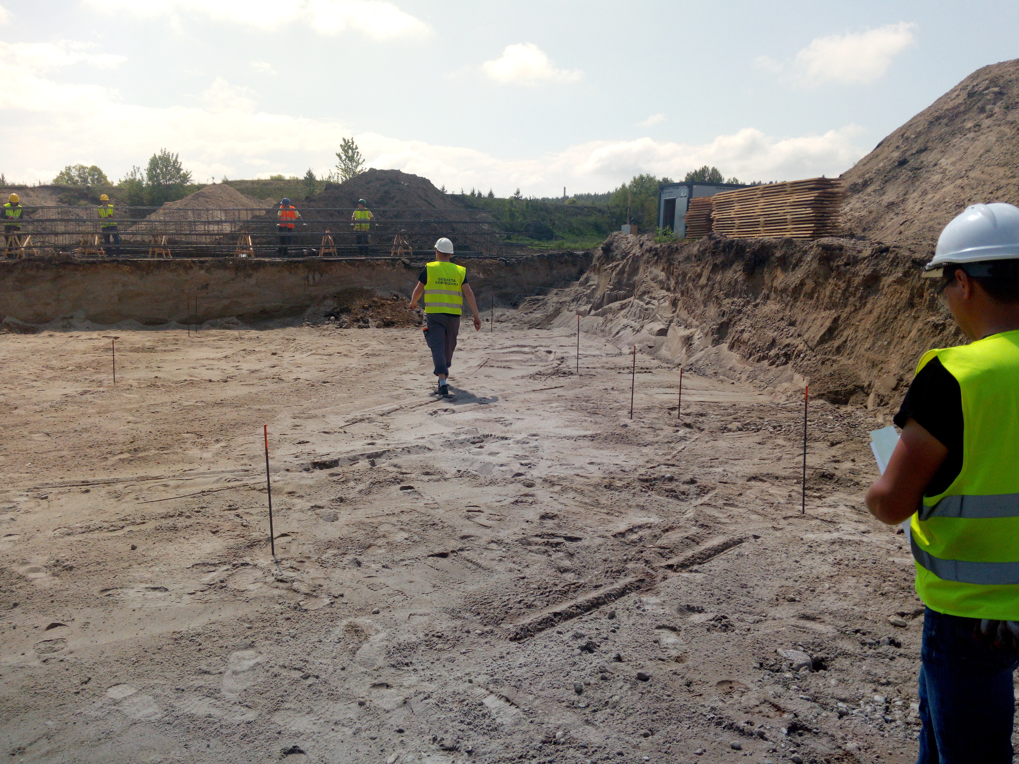 Przygotowanie terenu pod budowę: wykop z wyznaczonym obrysem fundamentów, pracownicy w odblaskowych kamizelkach i paliki geodezyjne. W tle składowane drewno.