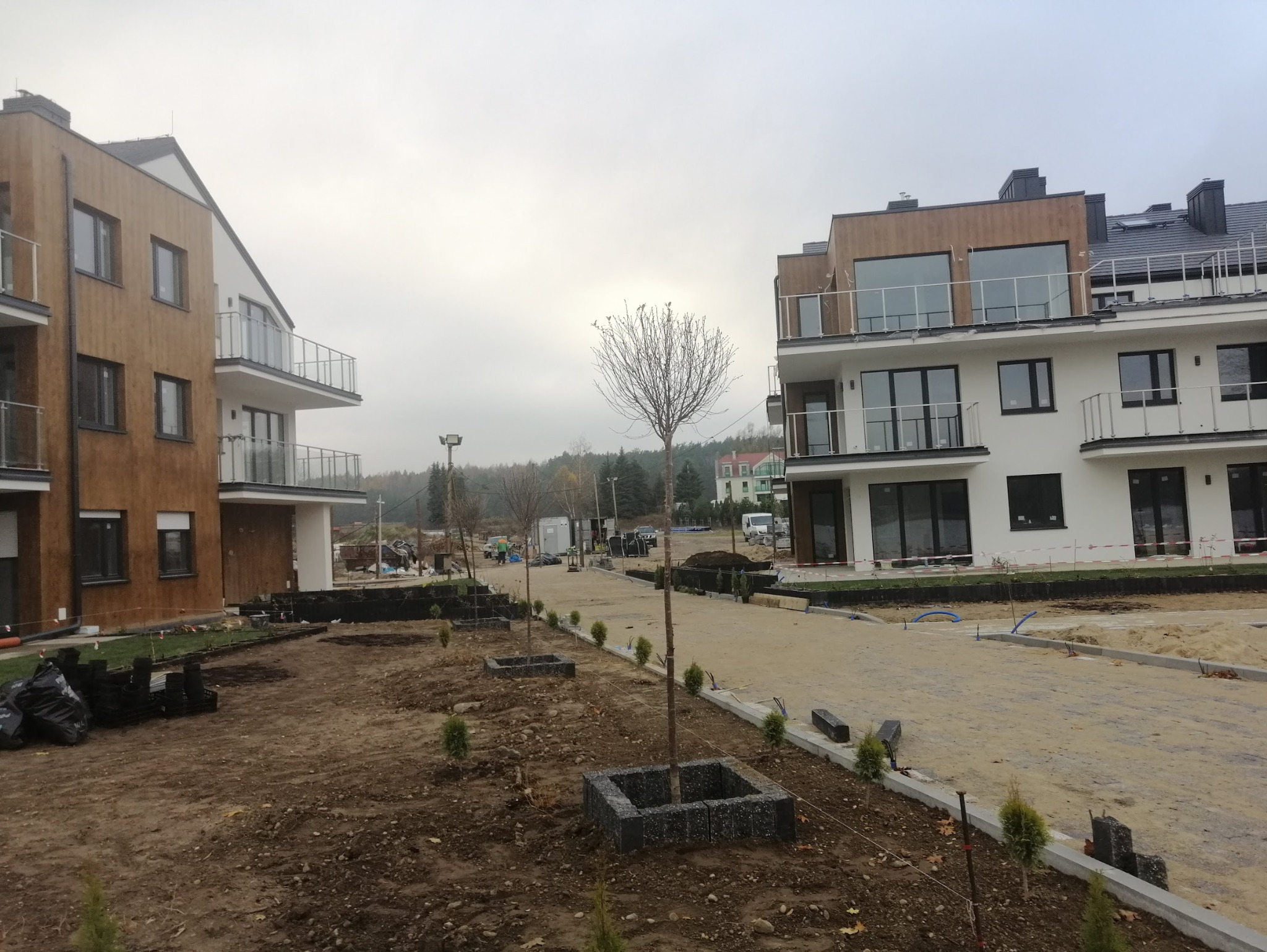 Nowoczesne budynki wielorodzinne z balkonami i tarasami, w trakcie zagospodarowania terenu wokół, z nasadzeniami drzew i krzewów. Brązowo-biała elewacja.