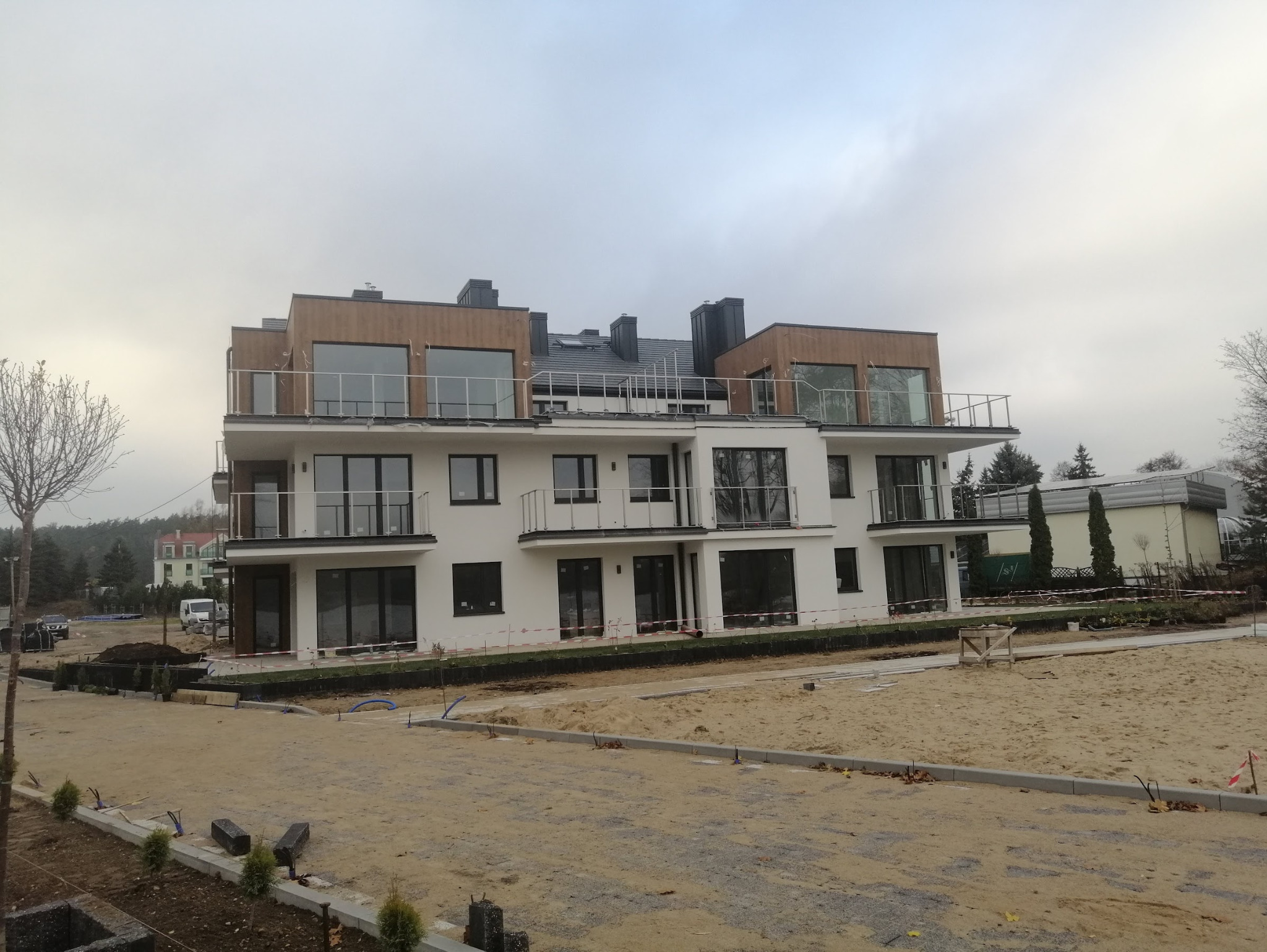 Nowoczesny, trzypiętrowy budynek z balkonami i dużymi oknami. Biała elewacja z drewnianymi akcentami. Teren wokół w trakcie prac wykończeniowych.