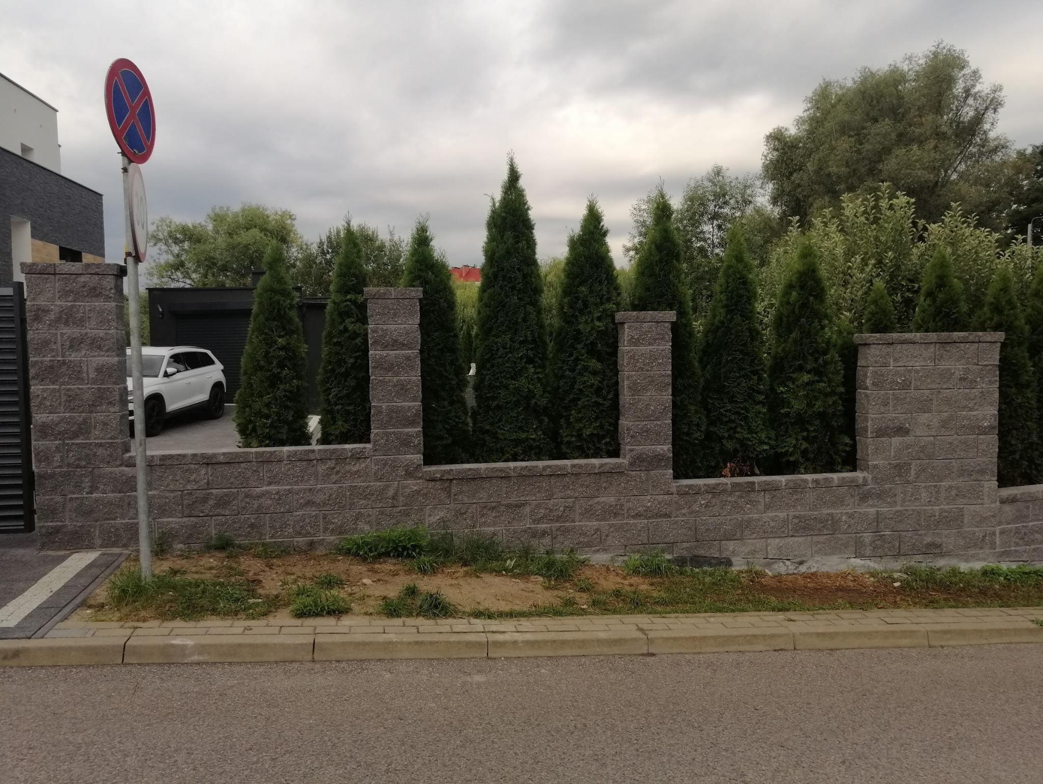 Nowoczesny mur z szarej cegły z wkomponowanymi tujami, tworzący ogrodzenie posesji z białym autem w tle. Widoczny znak drogowy. Umiarkowane oświetlenie.