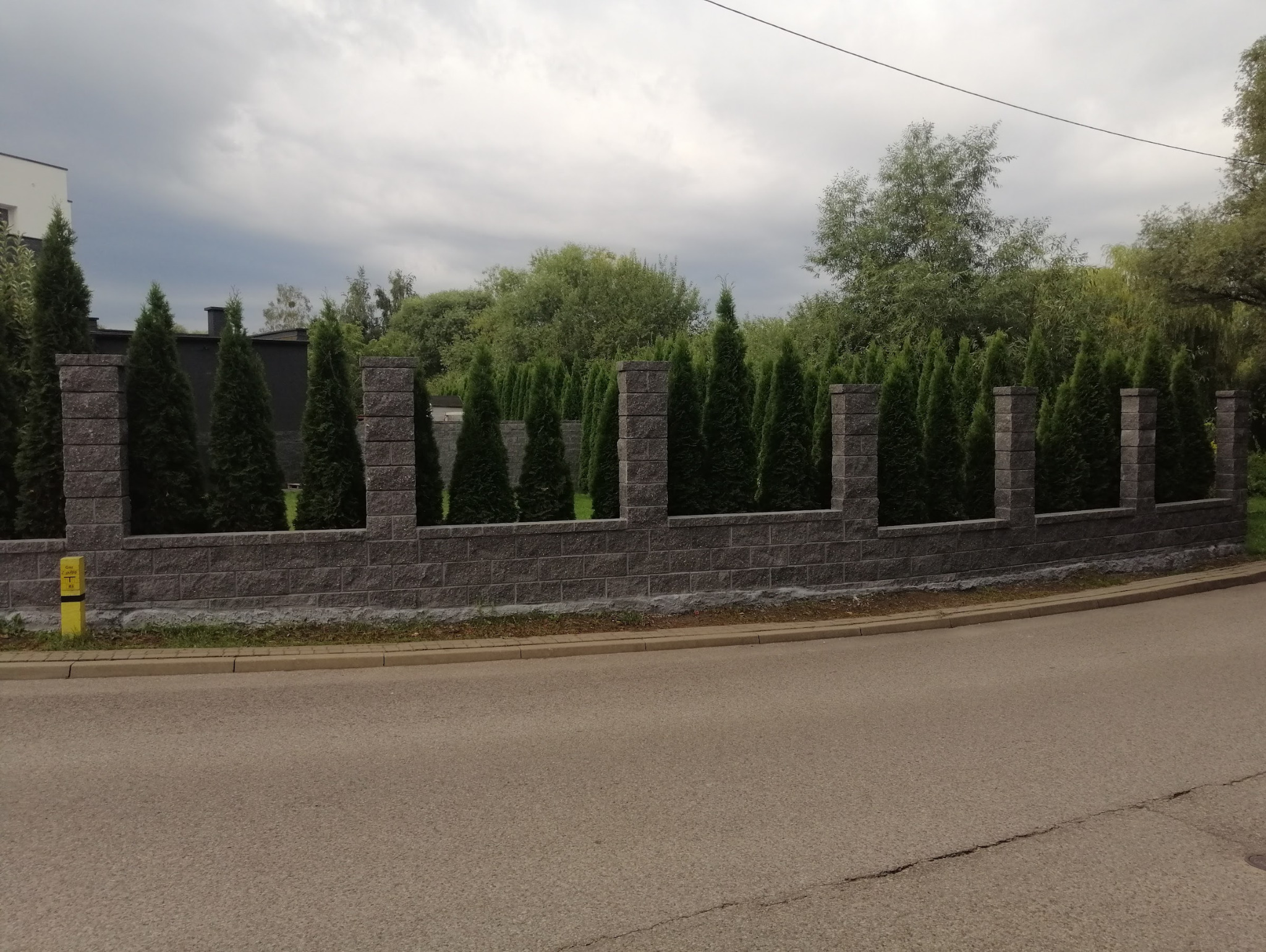 Nowoczesne ogrodzenie z szarej kostki łupanej, przerywane kolumnami, z zielonymi tujami w tle. Widok z ulicy, pochmurne niebo.
