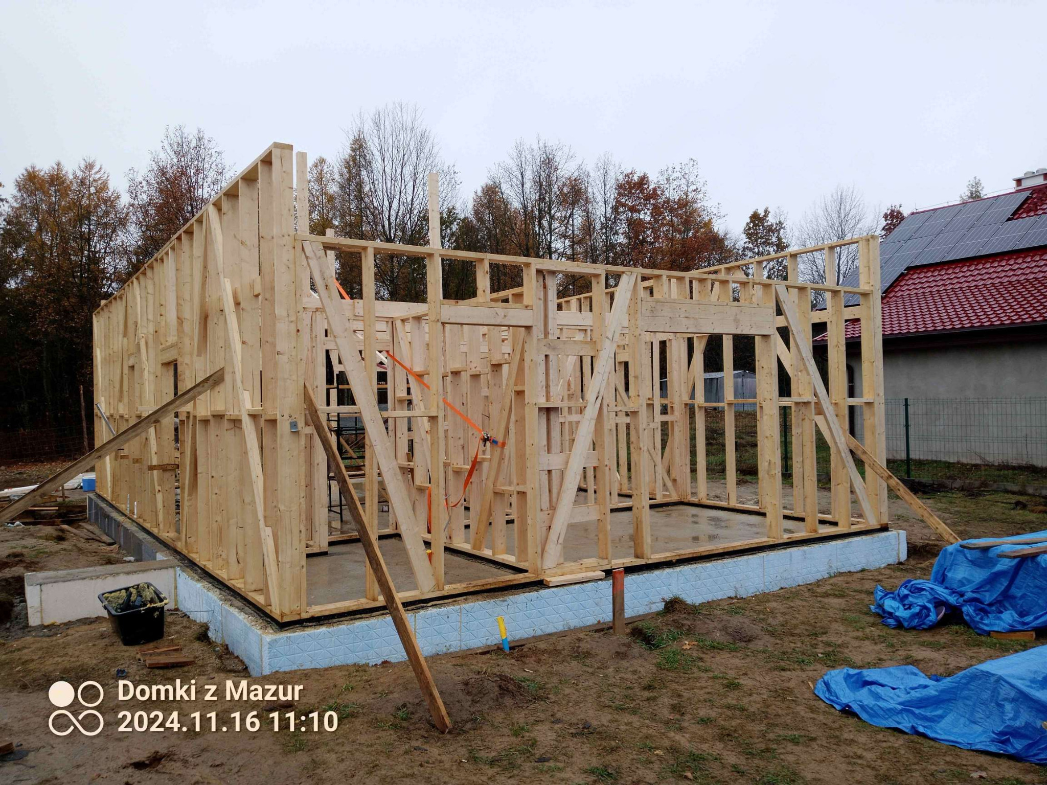 Szkieletowa konstrukcja domu w trakcie budowy, widoczne drewniane elementy ścian i fundamenty z izolacją, na zewnątrz rozłożona folia budowlana.