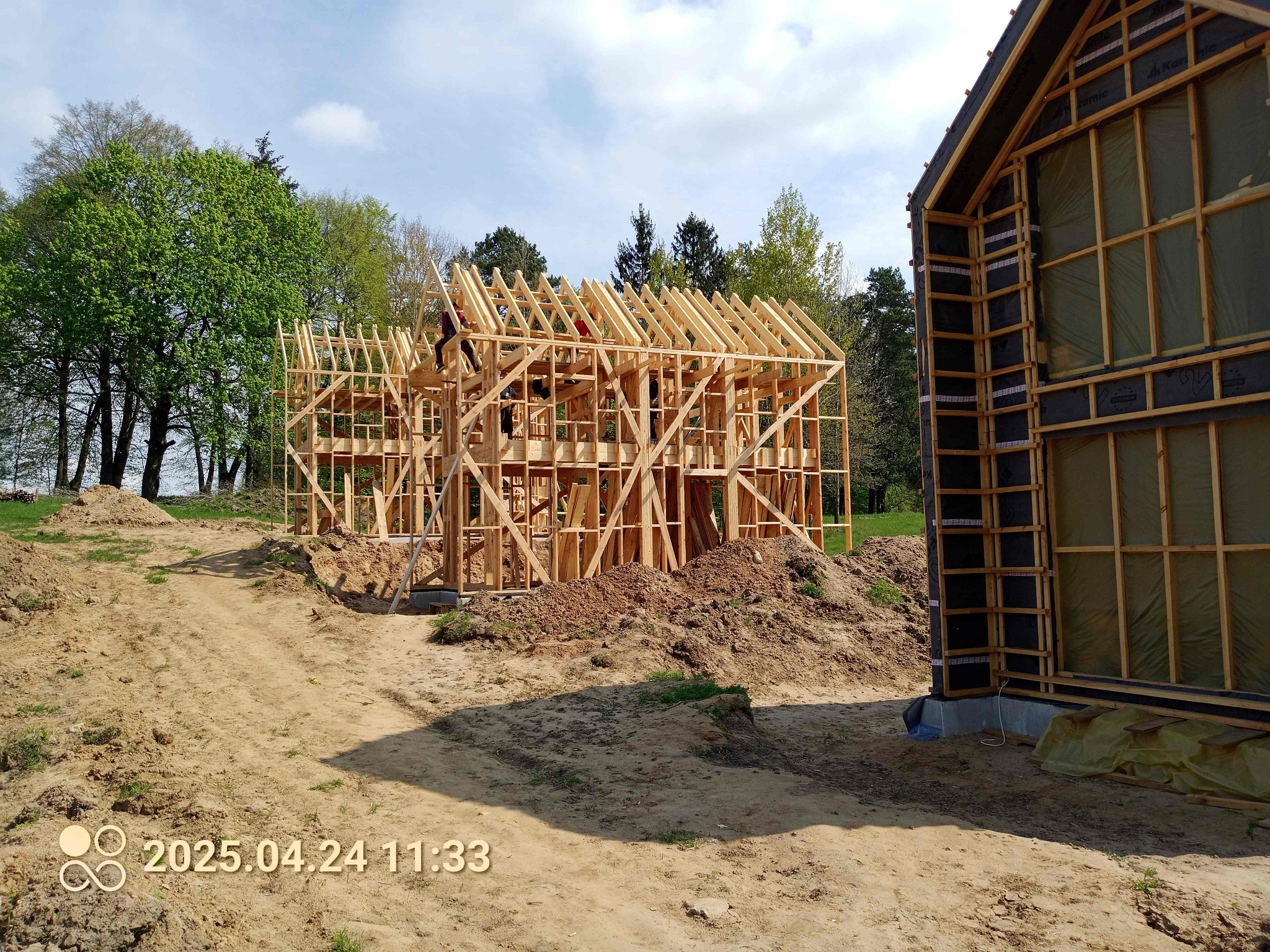 Wznoszenie szkieletu domu z drewna na działce. Widoczne elementy konstrukcyjne i pracownicy na dachu. Częściowo ocieplona ściana drugiego budynku w tle.