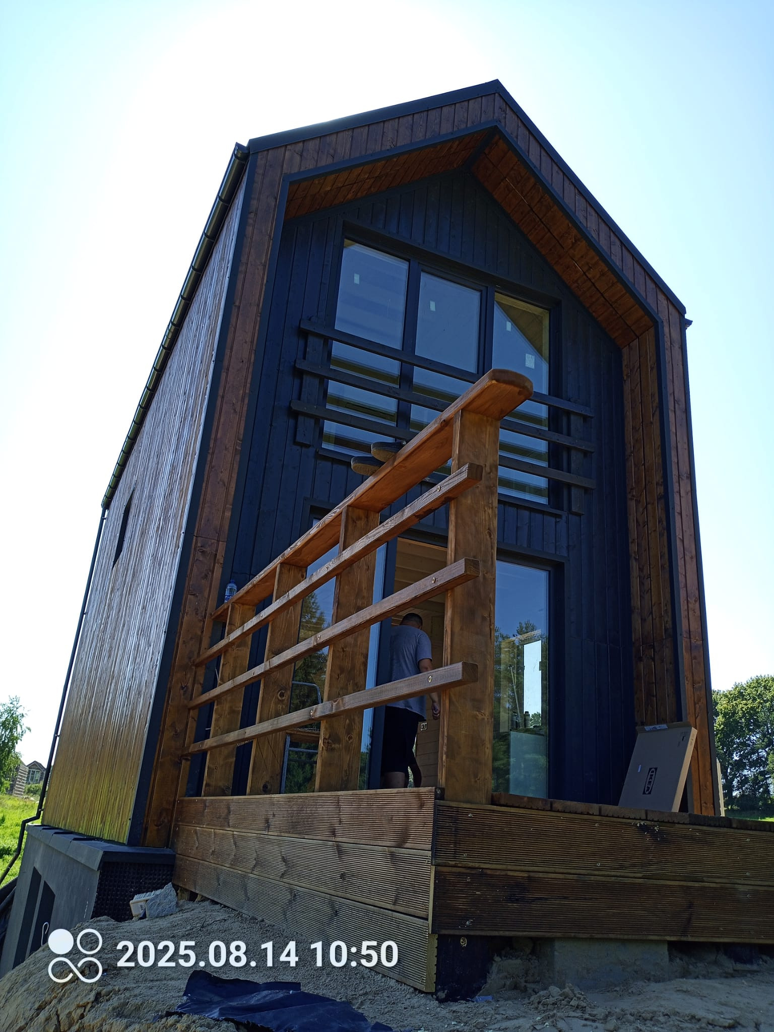 Nowoczesny dom szkieletowy z drewnianym tarasem i balustradą, widok z dołu, błękitne niebo w tle. Osoba na tarasie. Wykończenie w ciemnych kolorach.