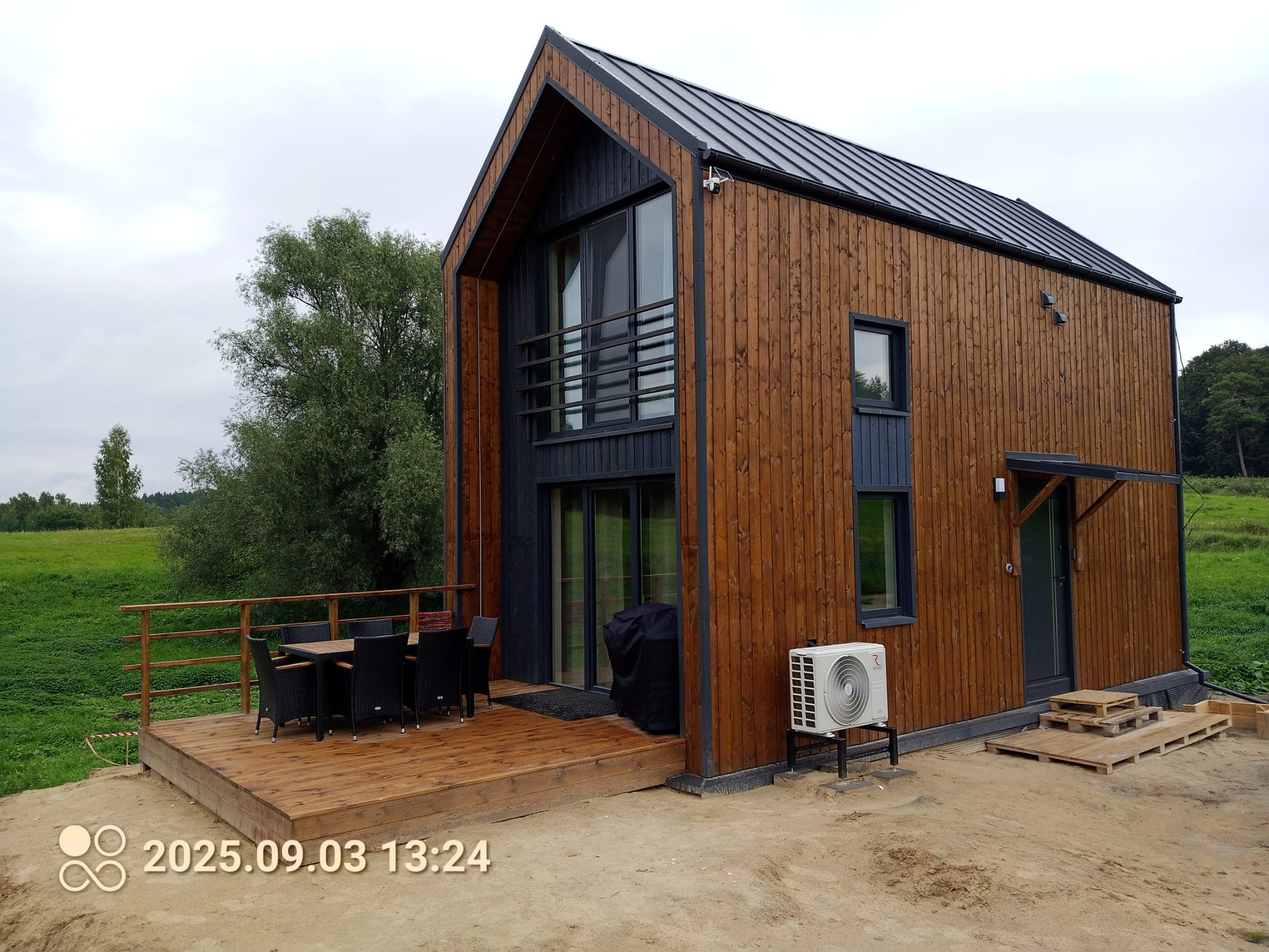 Nowoczesny dom modułowy z drewnianą elewacją, tarasem i klimatyzacją. Widok z zewnątrz w pochmurny dzień. Na pierwszym planie piasek i data na zdjęciu.