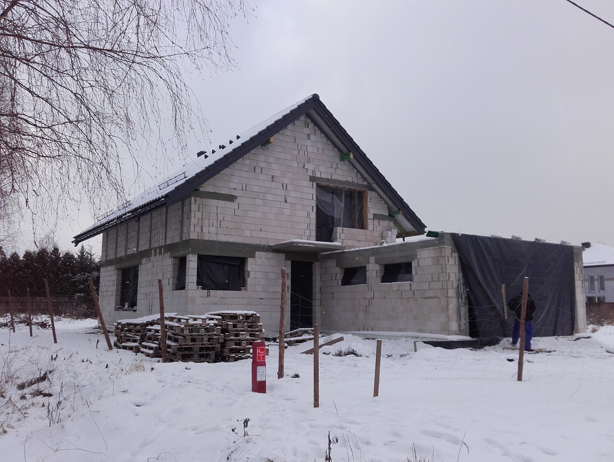 Budowa domu z betonu komórkowego w zimowej scenerii. Widoczne palety z materiałem i ogrodzenie z drewnianych słupków. Część okien zabezpieczona folią.