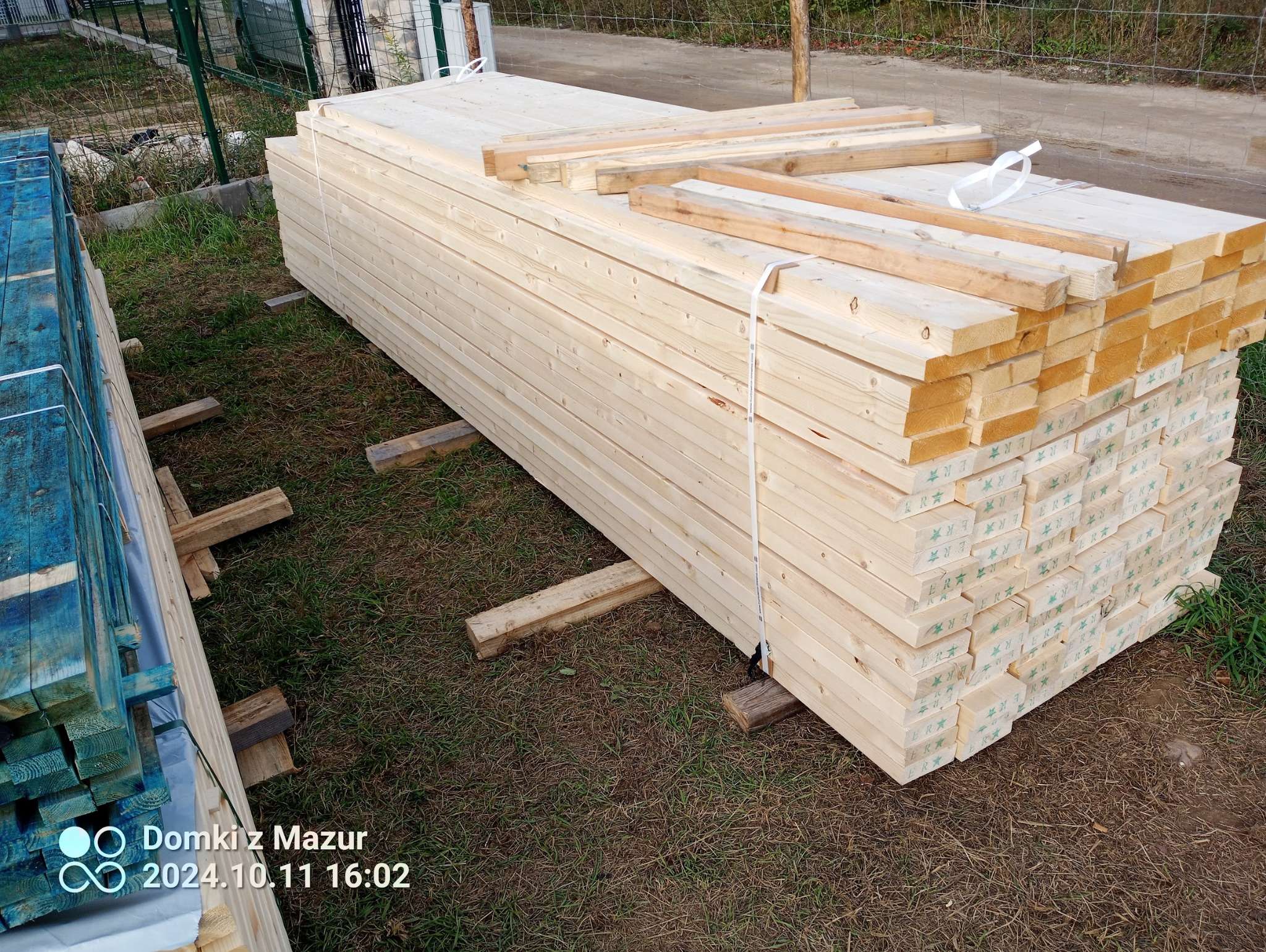 Stos jasnych, świeżych desek na trawie, zabezpieczonych drewnianymi belkami i białym sznurkiem. Część desek pomalowana na niebiesko. Widoczne zielone stemple na części desek.