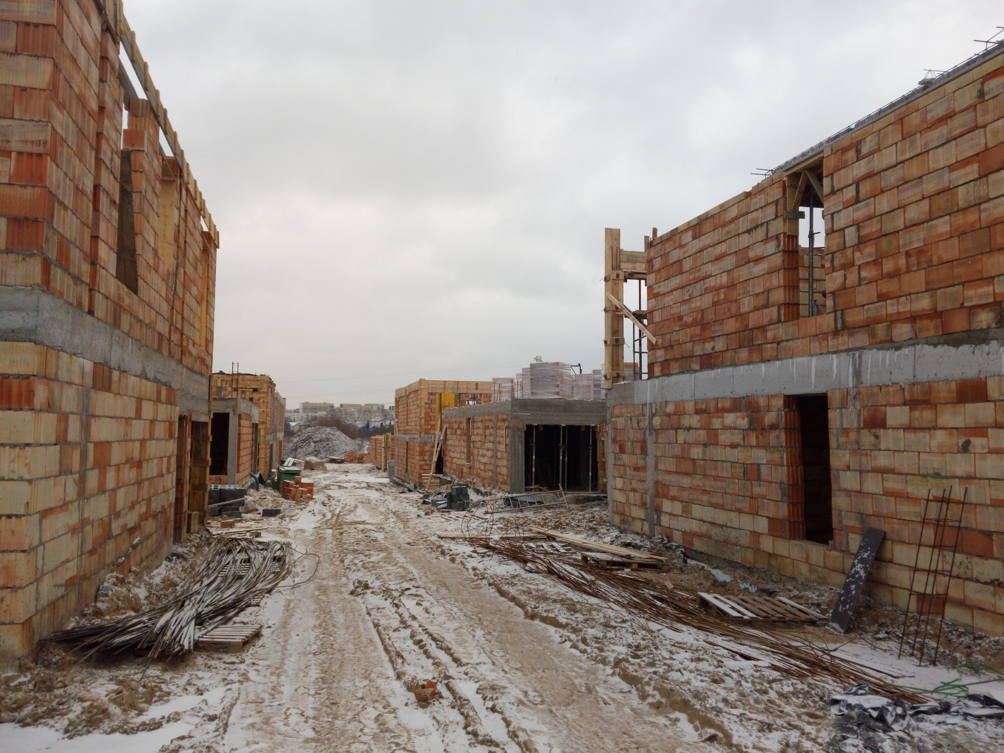 W zimowej scenerii, surowe mury z czerwonej cegły wznoszą się na osiedlu w budowie. Widoczny etap murowania ścian, z elementami szalunków i zbrojeń.
