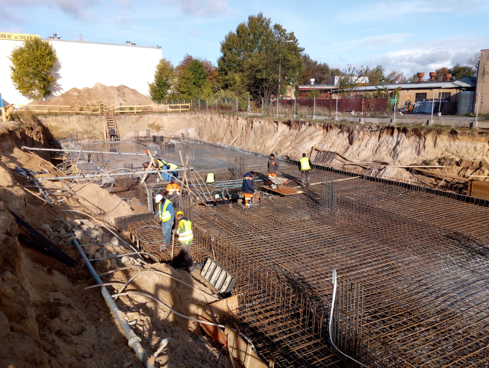 Zbrojenie fundamentów domu jednorodzinnego z widocznymi pracownikami w żółtych kamizelkach i kaskach, gotowe do zalania betonem. Wykop pod fundamenty.