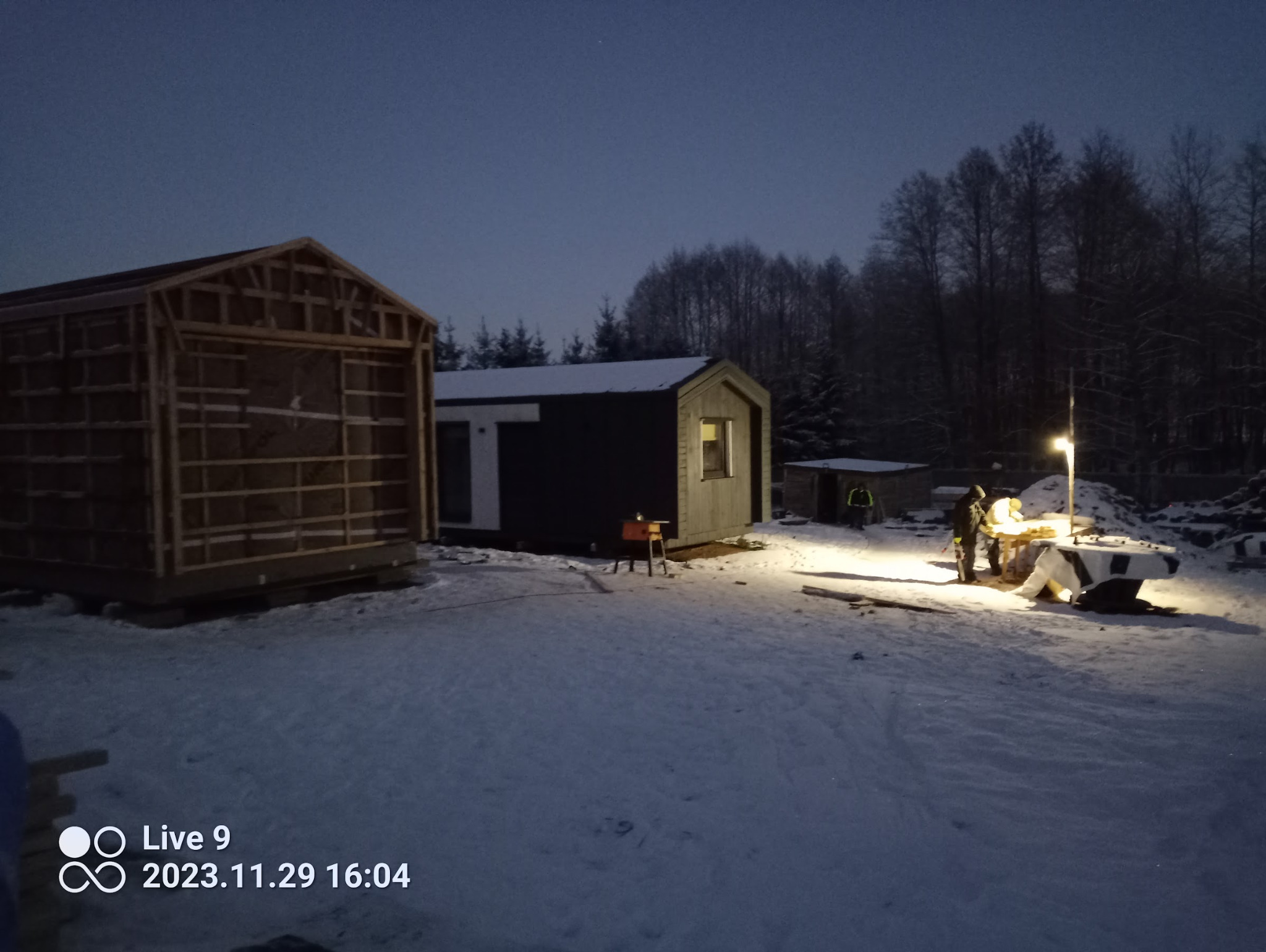 Budowa domu modułowego zimą: szkielet drewniany, gotowy moduł z oknem, pracownicy przy pracy, oświetlenie budowlane. Śnieg na ziemi i drzewa w tle.