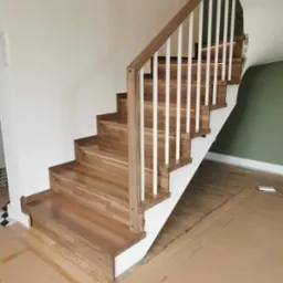 Jasne drewniane schody z białymi balustradami, widoczne zabezpieczenia podłogi kartonem, ściana w dwóch odcieniach.