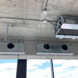 Jakub Nowak Luxtherm - Montaż systemu wentylacji mechanicznej z rekuperacją w surowym wnętrzu, widoczne kanały wentylacyjne pod sufitem betonowym, okablowanie i elementy montażowe, w tle widok na niebo przez okno.