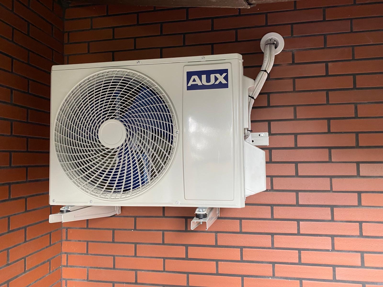 Zewnętrzna jednostka klimatyzacji marki AUX zamontowana na ceglanej ścianie, widoczne przewody w białej osłonie.