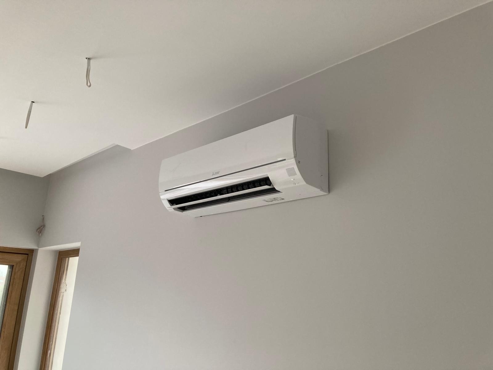 Biały klimatyzator Mitsubishi Electric zamontowany na szarej ścianie w jasnym pomieszczeniu z widocznymi przewodami na suficie.