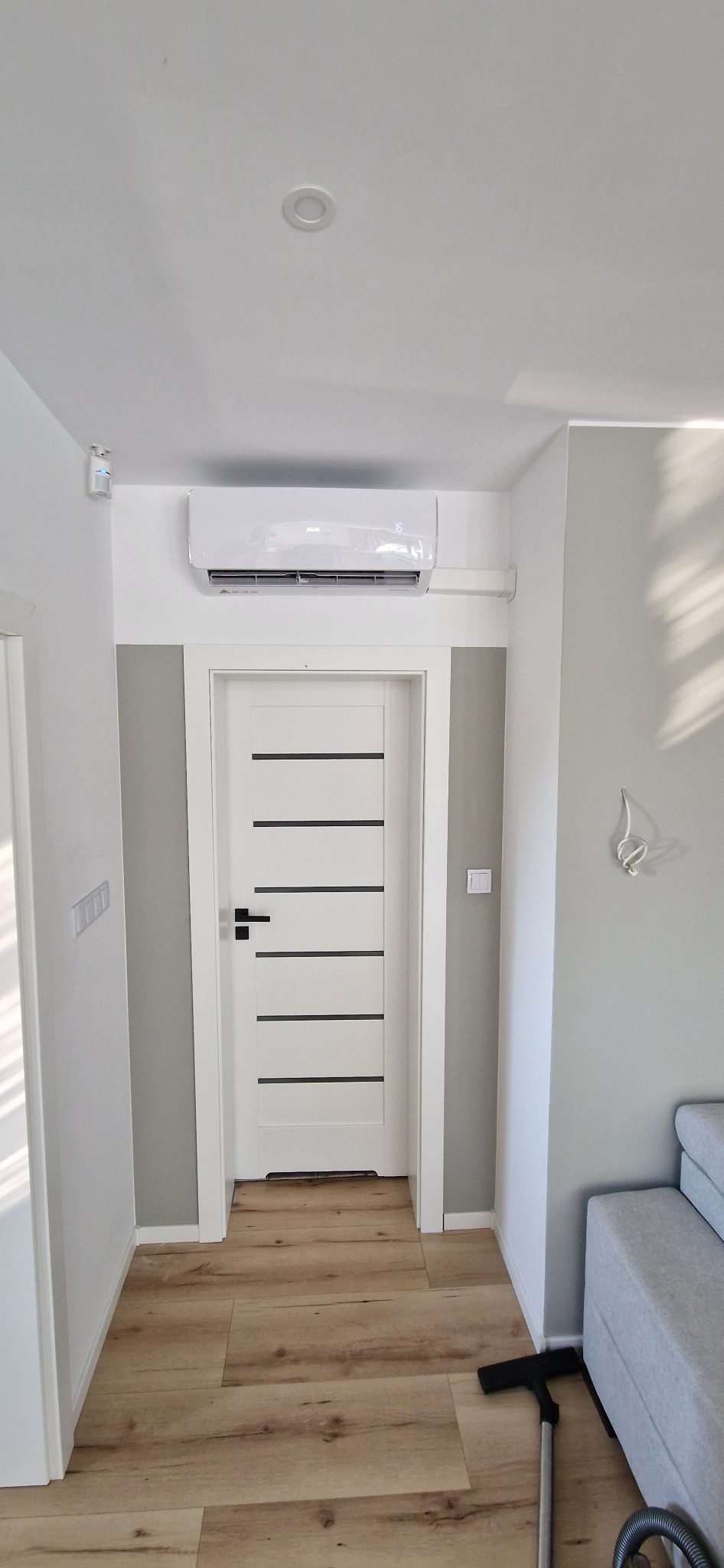 Biały klimatyzator ścienny zamontowany nad drzwiami w nowoczesnym mieszkaniu, widoczny fragment szarej sofy i odkurzacz na drewnianej podłodze.
