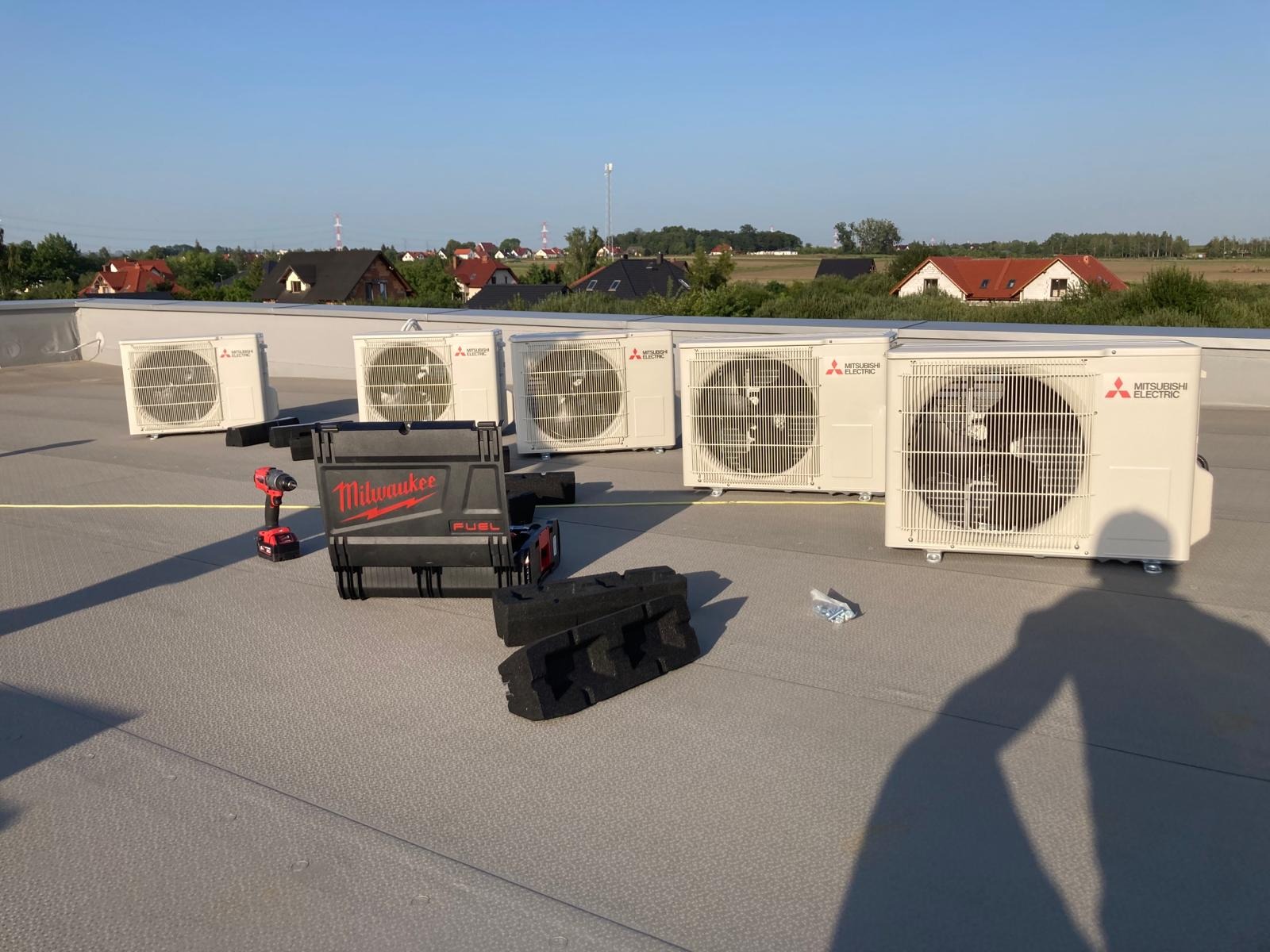 Białe jednostki zewnętrzne klimatyzacji Mitsubishi Electric ustawione na płaskim dachu, widoczna skrzynka narzędziowa Milwaukee FUEL, w tle domy jednorodzinne i błękitne niebo.