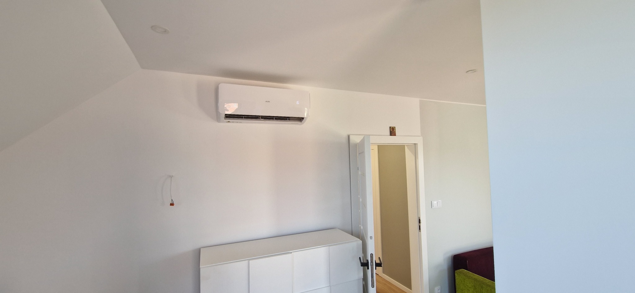 Biały klimatyzator AUX zamontowany na ścianie w jasnym pokoju z białymi meblami i otwartymi drzwiami.