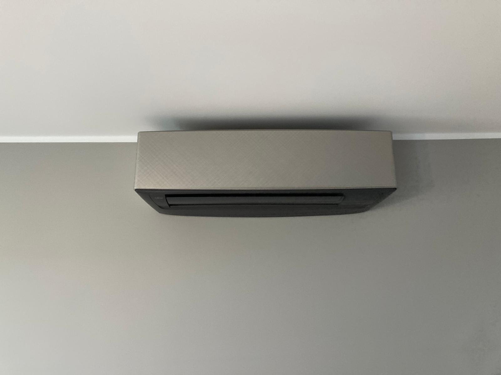 Szara jednostka wewnętrzna klimatyzatora, zamontowana na szarej ścianie pod białym sufitem, widoczna z dołu.