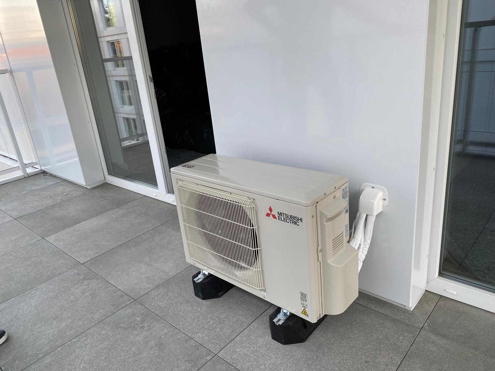Zewnętrzna jednostka klimatyzacji Mitsubishi Electric zamontowana na balkonie z szarymi płytkami, widoczne przewody i czarne podstawki antywibracyjne.