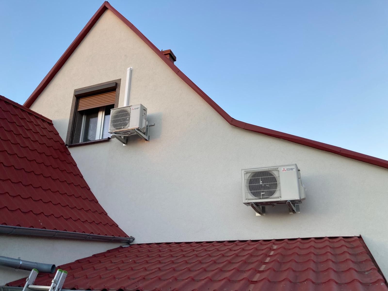 Dwa zewnętrzne moduły klimatyzacji marki Mitsubishi zamontowane na elewacji domu z czerwoną dachówką, jeden obok okna z roletami, drugi pod okapem dachu.