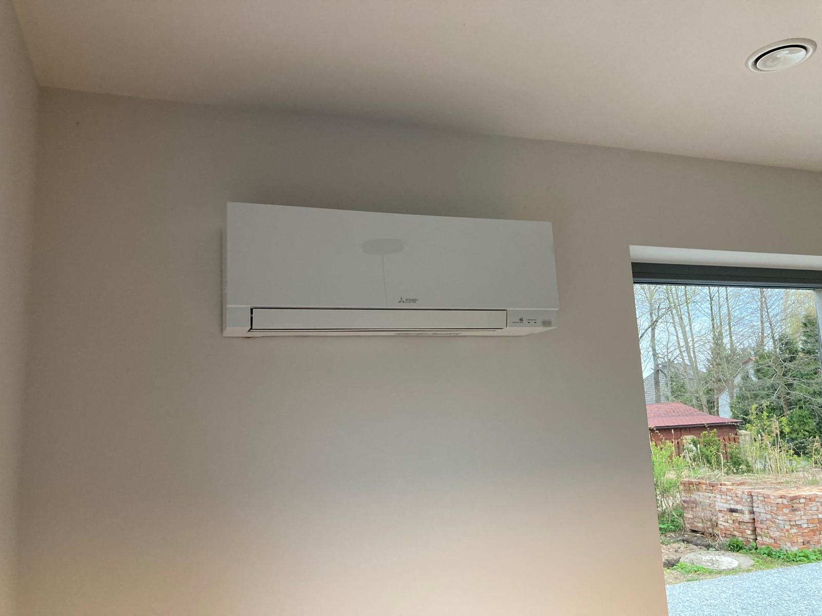 Biały klimatyzator marki Mitsubishi zamontowany na jasnej ścianie w pomieszczeniu, widoczne okno z widokiem na zieleń i cegły na zewnątrz.