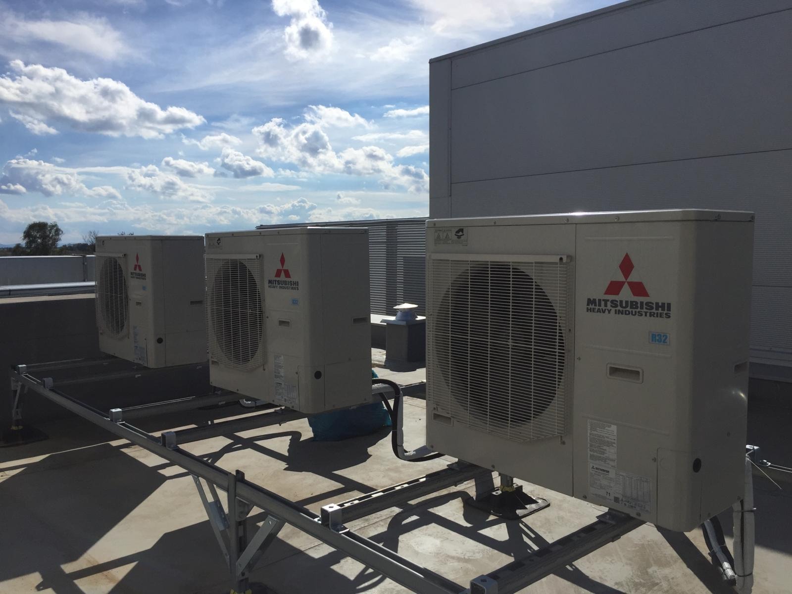 Trzy jednostki zewnętrzne klimatyzacji Mitsubishi Heavy Industries zamontowane na metalowej konstrukcji na dachu budynku przemysłowego, błękitne niebo z chmurami w tle.