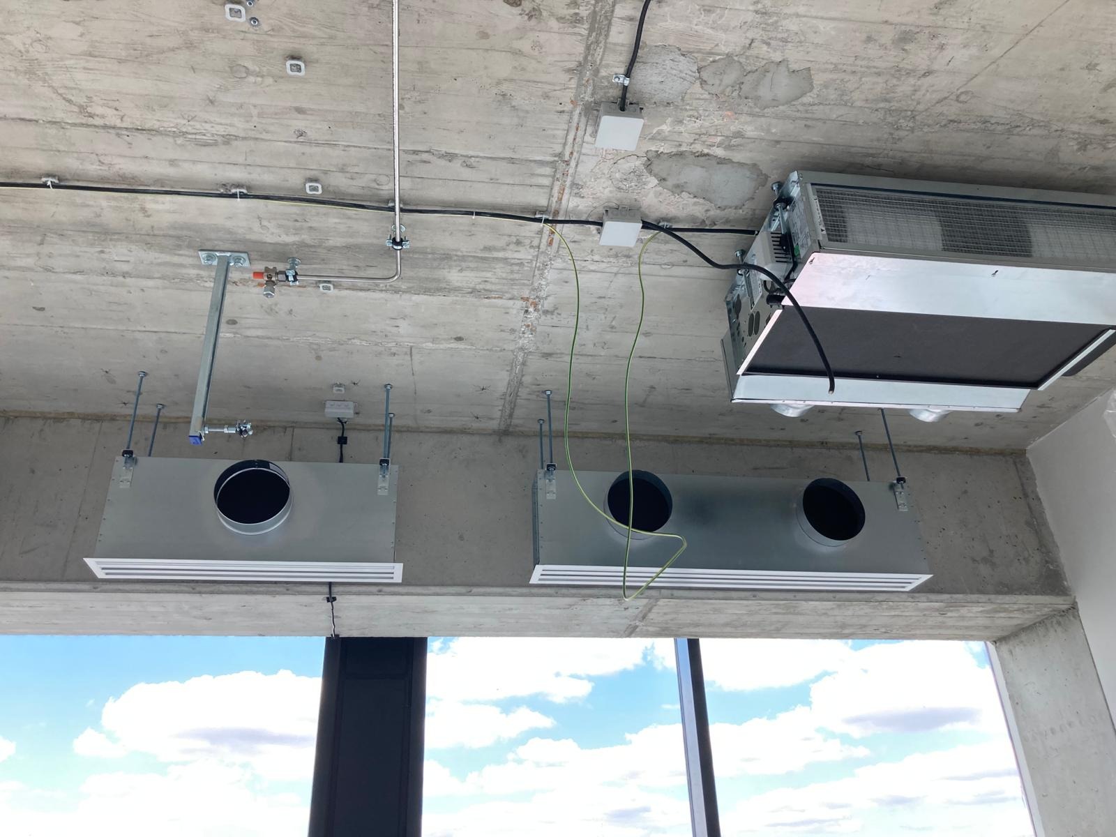 Montaż systemu wentylacji mechanicznej z rekuperacją w surowym wnętrzu, widoczne kanały wentylacyjne pod sufitem betonowym, okablowanie i elementy montażowe, w tle widok na niebo przez okno.