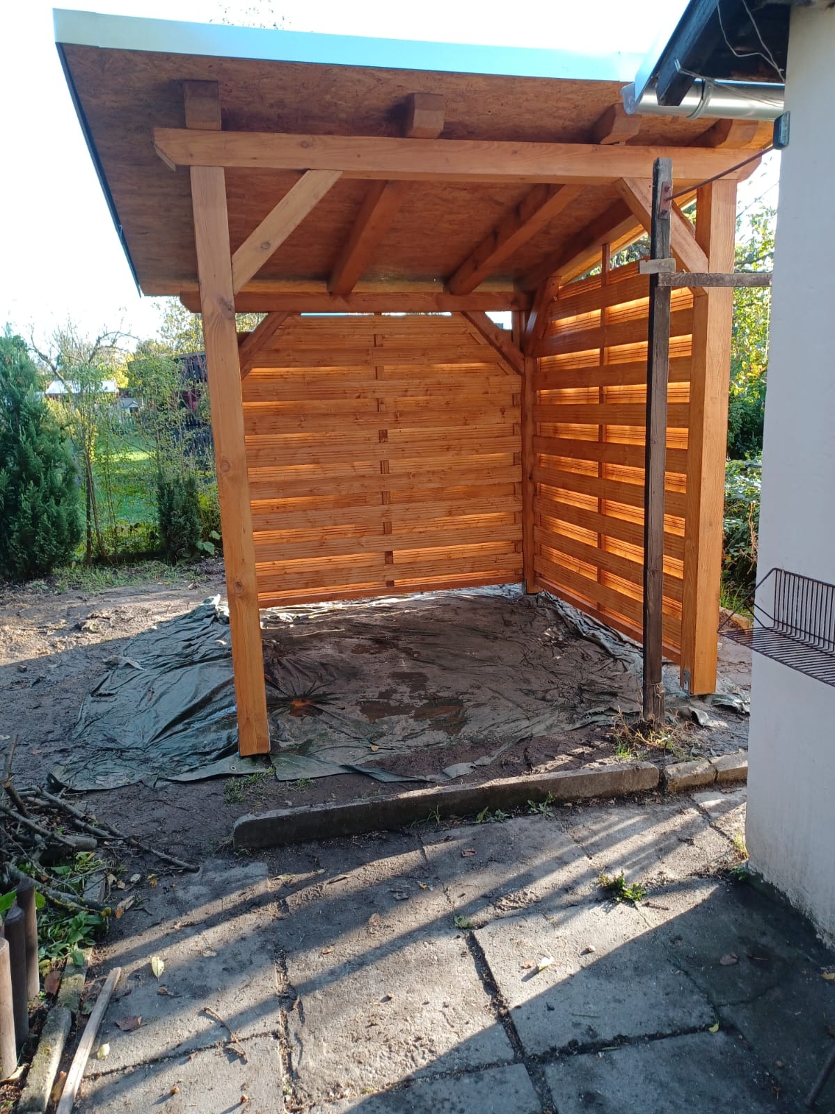 Drewniana konstrukcja zadaszenia w ogrodzie, częściowo osłonięta drewnianą kratą, na ziemi rozłożona plandeka, widoczne narzędzia i resztki materiałów.