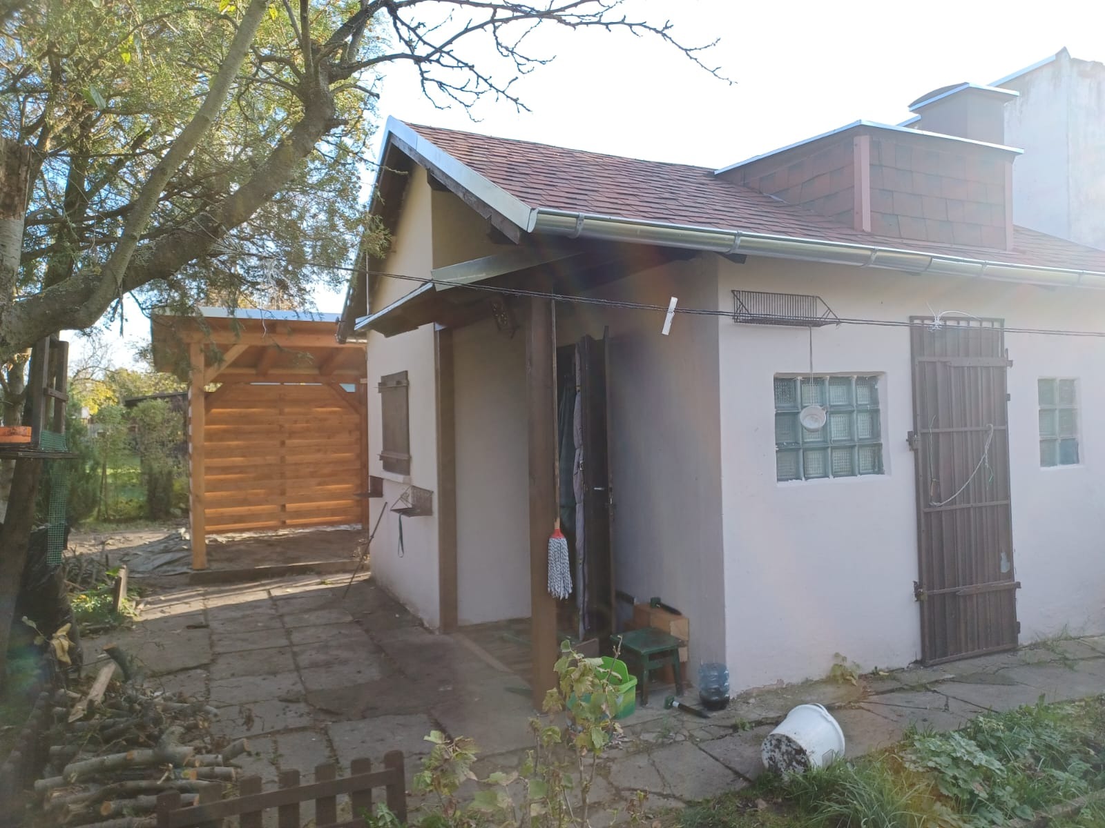 Niski budynek gospodarczy z drewnianą wiatą w tle, widoczne narzędzia i akcesoria porządkowe przy wejściu.