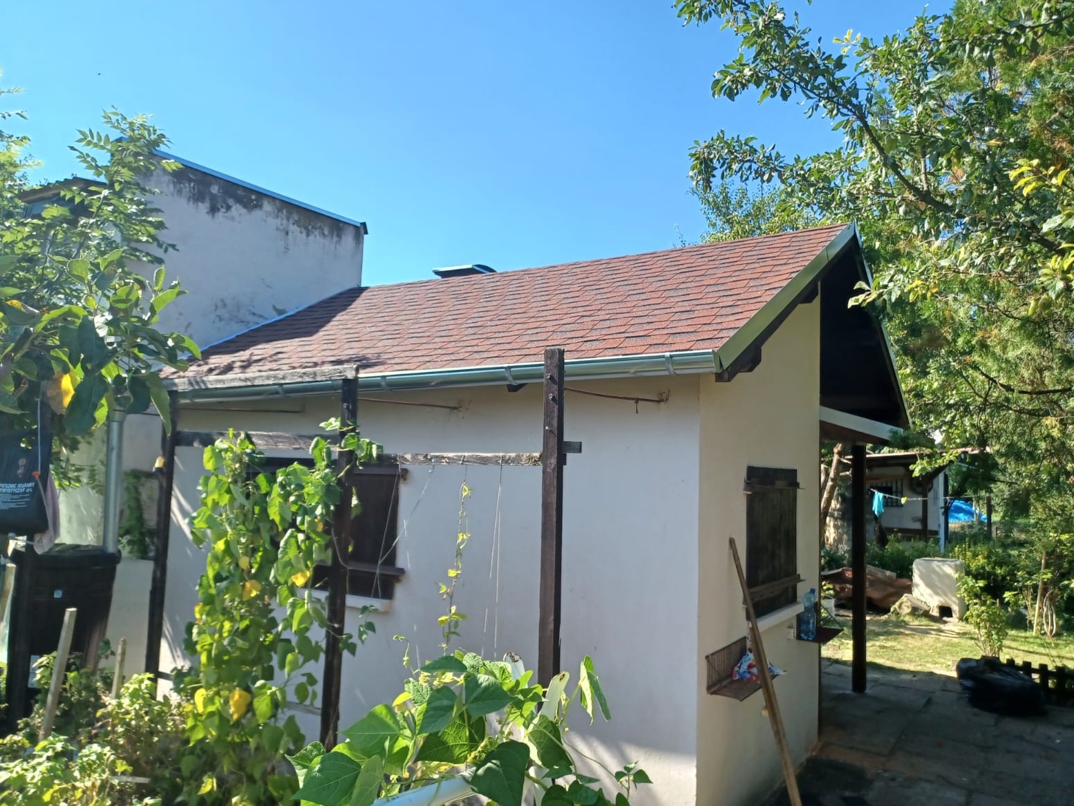 Niski budynek gospodarczy z nowym, brązowym dachem z gontu bitumicznego, widoczne orynnowanie, drewniane okiennice i bujna roślinność w tle.