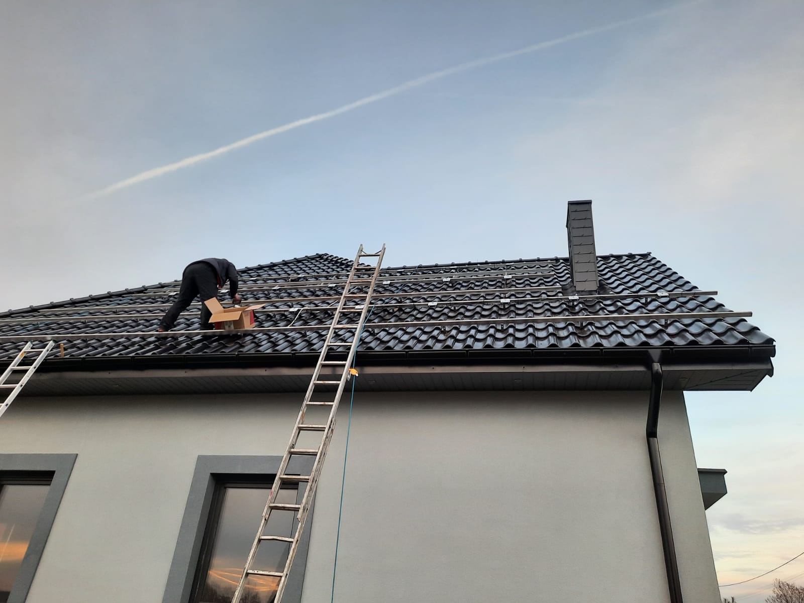 Montaż konstrukcji pod panele słoneczne na dachu z czarnej dachówki karpiówki, widoczne drabiny opierające się o ścianę budynku.