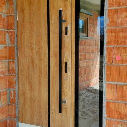 F.H.U RES DOM TOMASZ ZIARNIK - Nowoczesne drzwi zewnętrzne z drewnopodobnym panelem i czarnymi uchwytami, zamontowane w surowym murze z cegły, z widocznym fragmentem okna w odbiciu szyby bocznej.