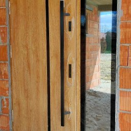 F.H.U RES DOM TOMASZ ZIARNIK - Nowoczesne drzwi wejściowe z drewnopodobnym wykończeniem i czarnym, długim uchwytem, zamontowane w surowym murze, z widocznym oknem bocznym odbijającym krajobraz zewnątrz.