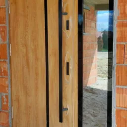 Nowoczesne drzwi wejściowe z drewnopodobnym wykończeniem i czarnym, długim uchwytem, zamontowane w surowym murze, z widocznym oknem bocznym odbijającym krajobraz zewnątrz.