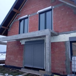 F.H.U RES DOM TOMASZ ZIARNIK - Surowy stan budowy domu jednorodzinnego z zamontowanymi oknami i zewnętrznymi roletami w kolorze szarym, widoczne elementy konstrukcyjne dachu i surowe mury z czerwonej cegły.