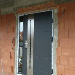 F.H.U RES DOM TOMASZ ZIARNIK - Montaż szarych drzwi zewnętrznych z pionowym, srebrnym panelem w surowym murze, widoczna pianka montażowa i paleta budowlana.