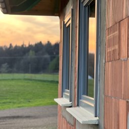 F.H.U RES DOM TOMASZ ZIARNIK - Dwa szare okna w surowej ścianie z czerwonej cegły, widok na zachodzące słońce i zieleń w tle, widać parapety zewnętrzne.