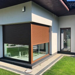 F.H.U RES DOM TOMASZ ZIARNIK - Narożne okno z brązowymi roletami zewnętrznymi, częściowo opuszczonymi, w nowoczesnym domu z białymi ścianami, widok z zewnątrz na zadbany trawnik i kostkę brukową