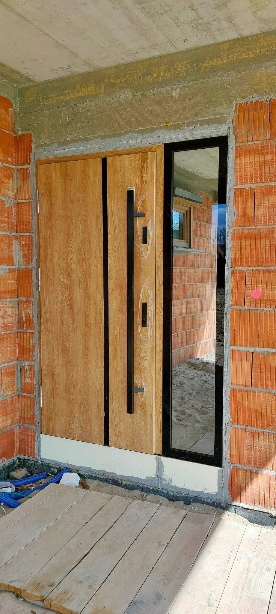 Nowoczesne drzwi zewnętrzne z drewnopodobnym panelem i czarnymi uchwytami, zamontowane w surowym murze z cegły, z widocznym fragmentem okna w odbiciu szyby bocznej.