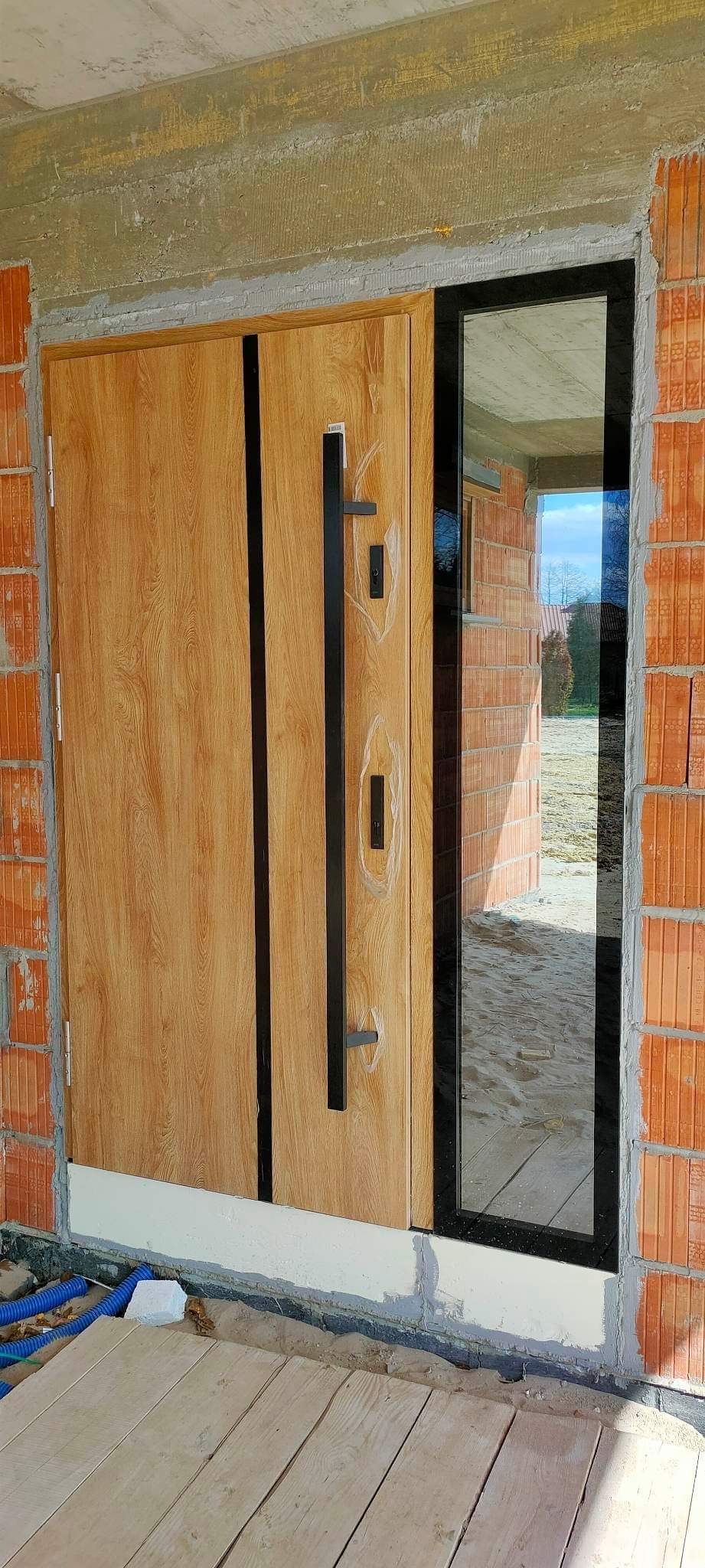 Nowoczesne drzwi wejściowe z drewnopodobnym wykończeniem i czarnym, długim uchwytem, zamontowane w surowym murze, z widocznym oknem bocznym odbijającym krajobraz zewnątrz.