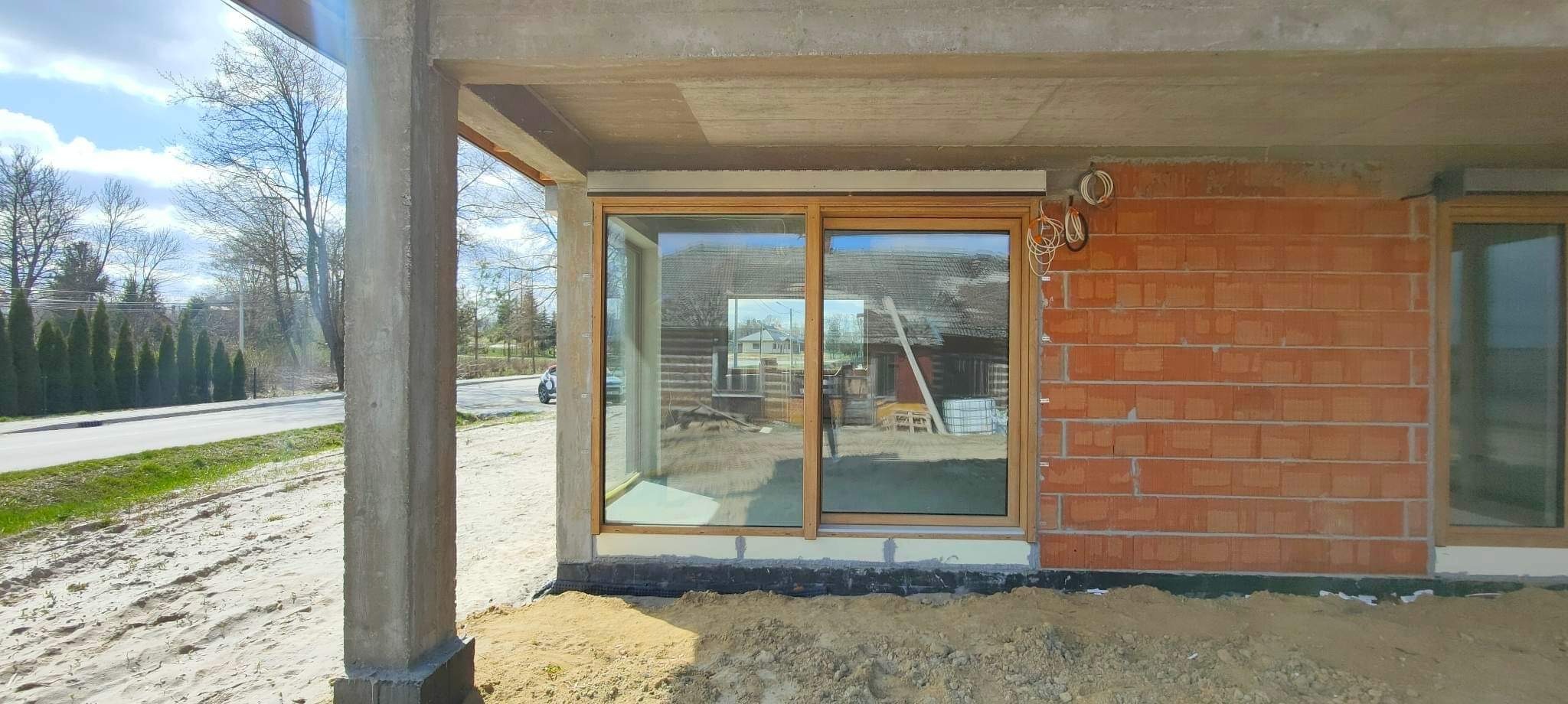 Montaż dużego okna przesuwnego z drewnianą ramą w surowym stanie budynku z cegły, widoczne przewody elektryczne i betonowe elementy konstrukcyjne.