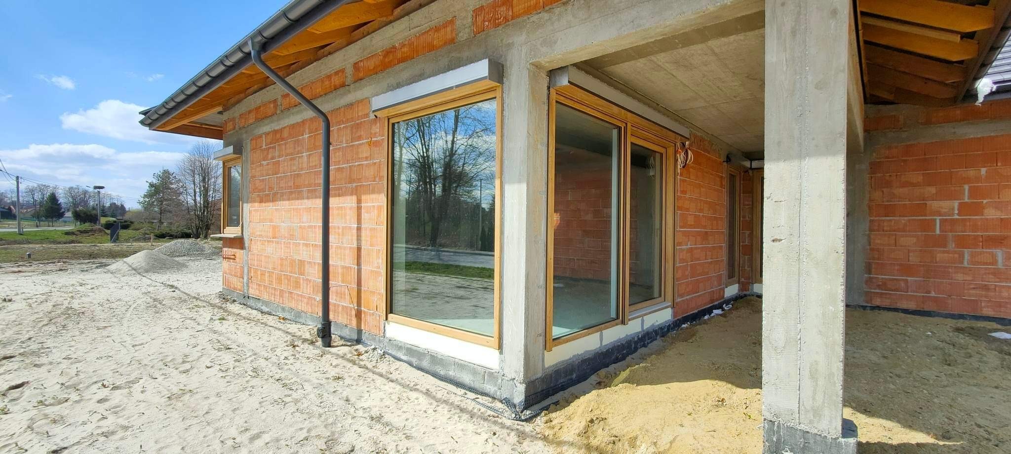 Surowy stan budynku w trakcie budowy, widoczne niezabudowane okna z drewnianymi ramami i szare rolety zewnętrzne, ściany z czerwonej cegły, betonowe filary i piasek wokół budynku.