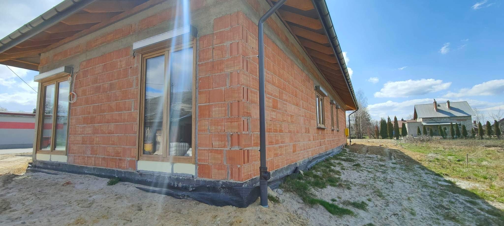 Surowy budynek jednorodzinny w trakcie budowy z zamontowanymi oknami z roletami zewnętrznymi, widoczne cegły dziurawki, folia fundamentowa i obróbka okien.