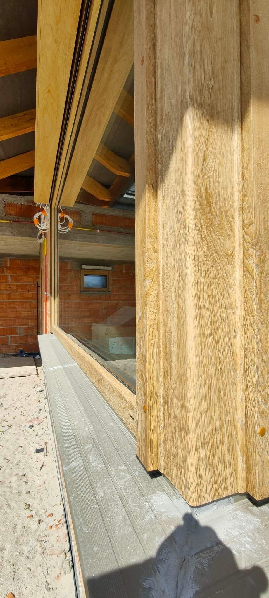 Fragment montażu okna przesuwnego z drewnianą ramą i grubym, drewnianym słupkiem, widoczny mur z cegły i elementy konstrukcyjne dachu w surowym stanie.