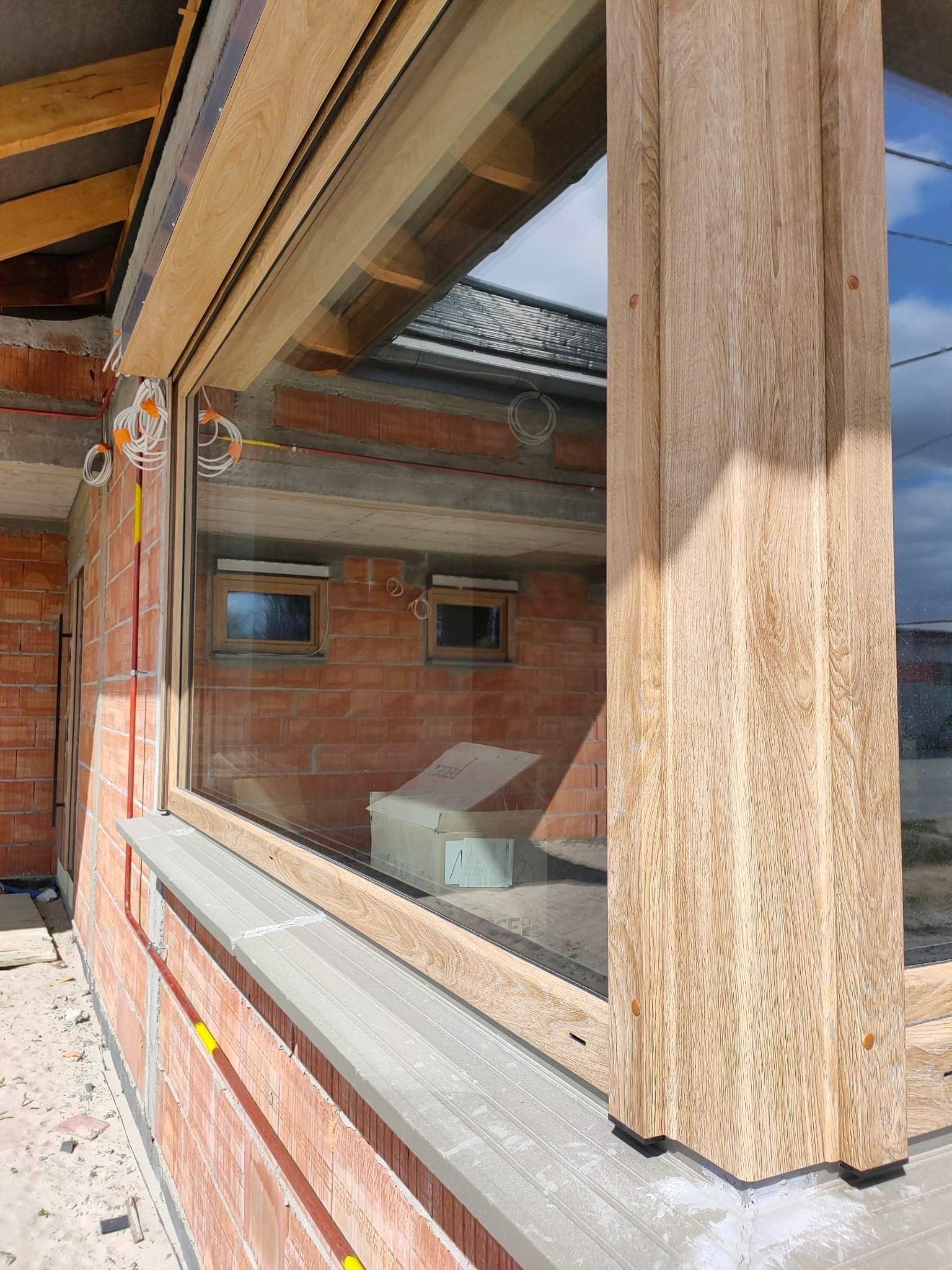 Montaż okna z drewnianą ramą w surowym stanie budynku z czerwonej cegły. Widoczne przewody elektryczne i elementy konstrukcyjne dachu.